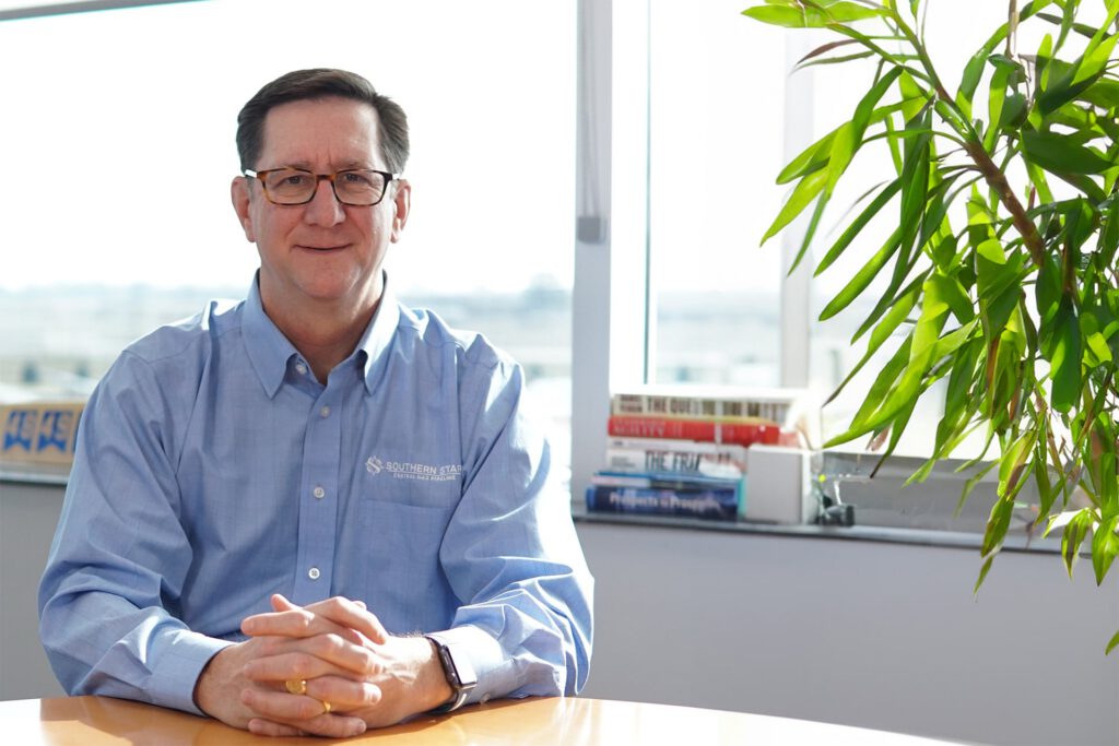 Man sitting at a desk in a blue button up shirt with a green plant on the right side.